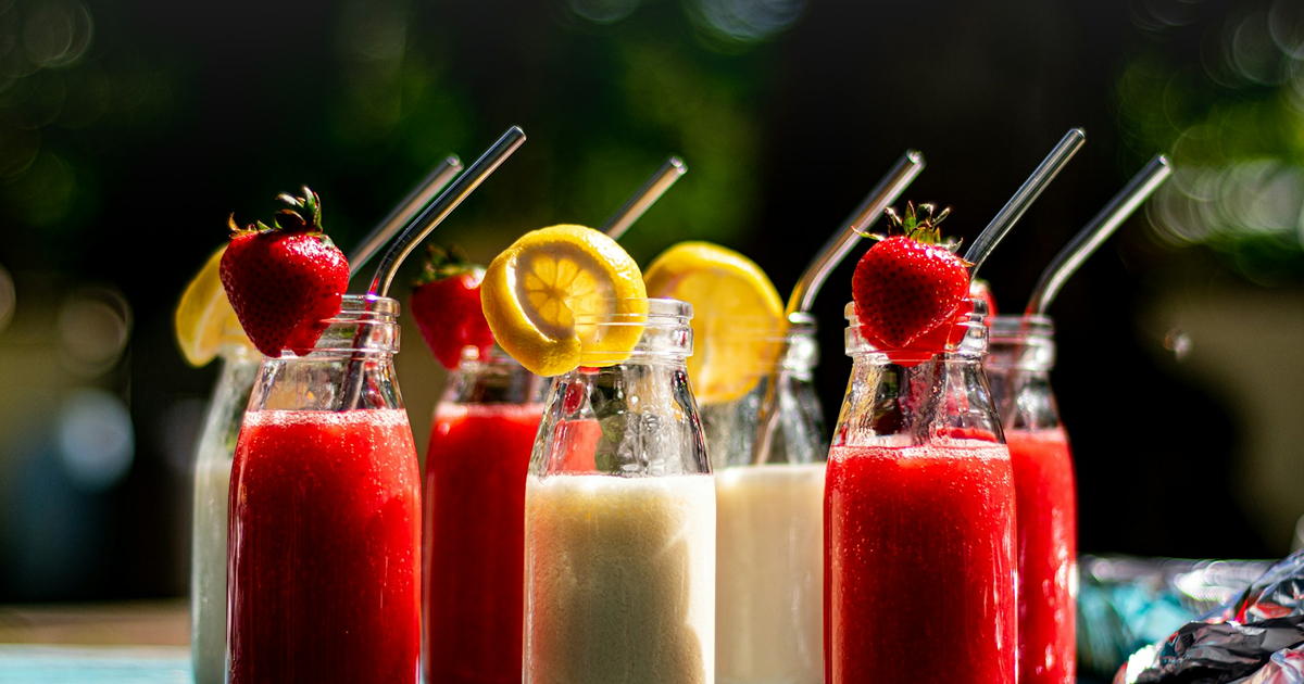 Glass bottles of fruit smoothies with metal straws, garnished with strawberries and lemon slices.